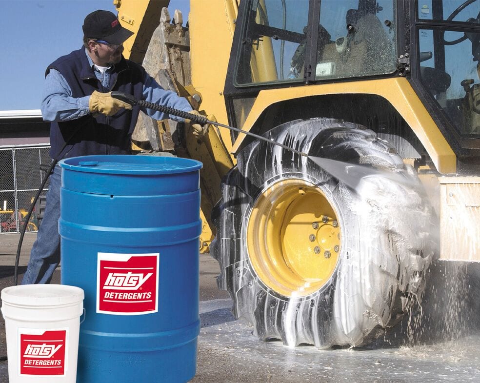 Large barrel and 5 gallon bucket of detergent in front of a man powerwashing a piece of large equipment